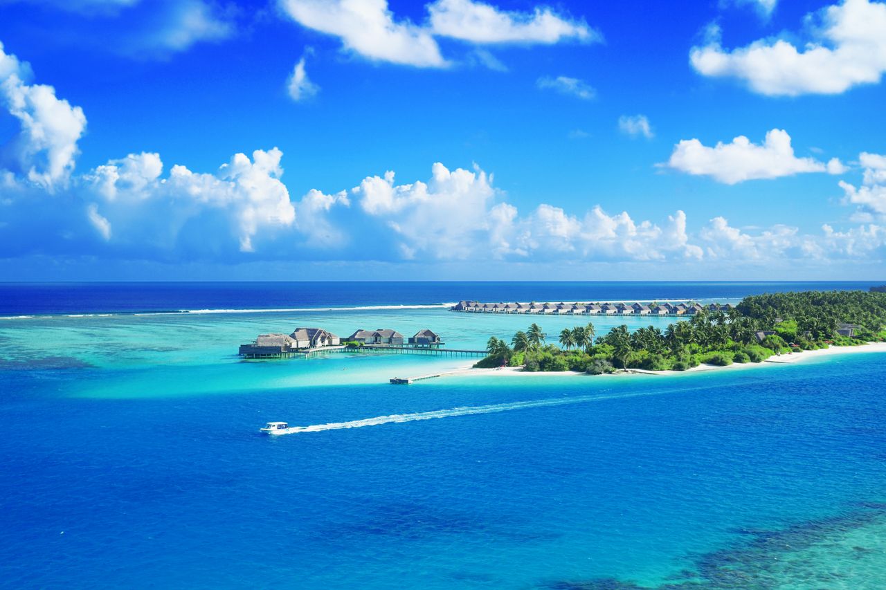 Home Crystal clear waters surround a luxurious Maldives resort under a bright blue sky.